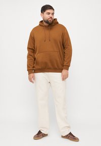 Brązowy bawełniany hoodie z kieszenią z przodu, kapturem z sznurkiem i ściągaczami na rękawach, w połączeniu z jasnymi kremowymi spodniami i brązowym obuwiem sportowym.
