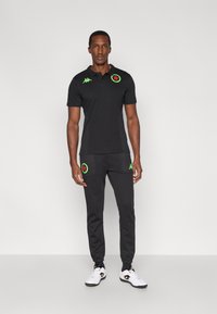 Čierne polo tričko so zelenými logami, v kombinácii s tmavými teplákmi so zodpovedajúcimi vreckovými logami. Obuv je biela s čiernymi detailmi.