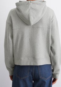 Osoba oblečená v světle šedé zkrácené mikině a tmavě modrých džínách, zobrazená zezadu na pozadí jednoduché bílé zdi.