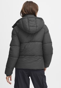 Dunjakke i mørkegrått med hette. Kviltet design, med en glatt tekstur og elastiske mansjetter. Snittet er litt løst for varme.