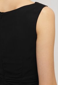 Vestido negro sin mangas con escote redondo, tejido suave y detalle fruncido en la cintura con una cremallera oculta en la parte trasera.