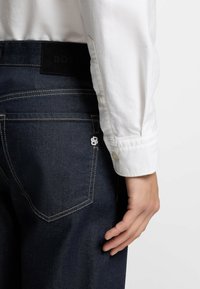 Tummansiniset farkut, joissa näkyvä takatasku, kultaiset ompeleet ja musta nahkalappu, jossa "BOSS" vyötäröllä. Valkoinen paidan kalvosin näkyvissä.