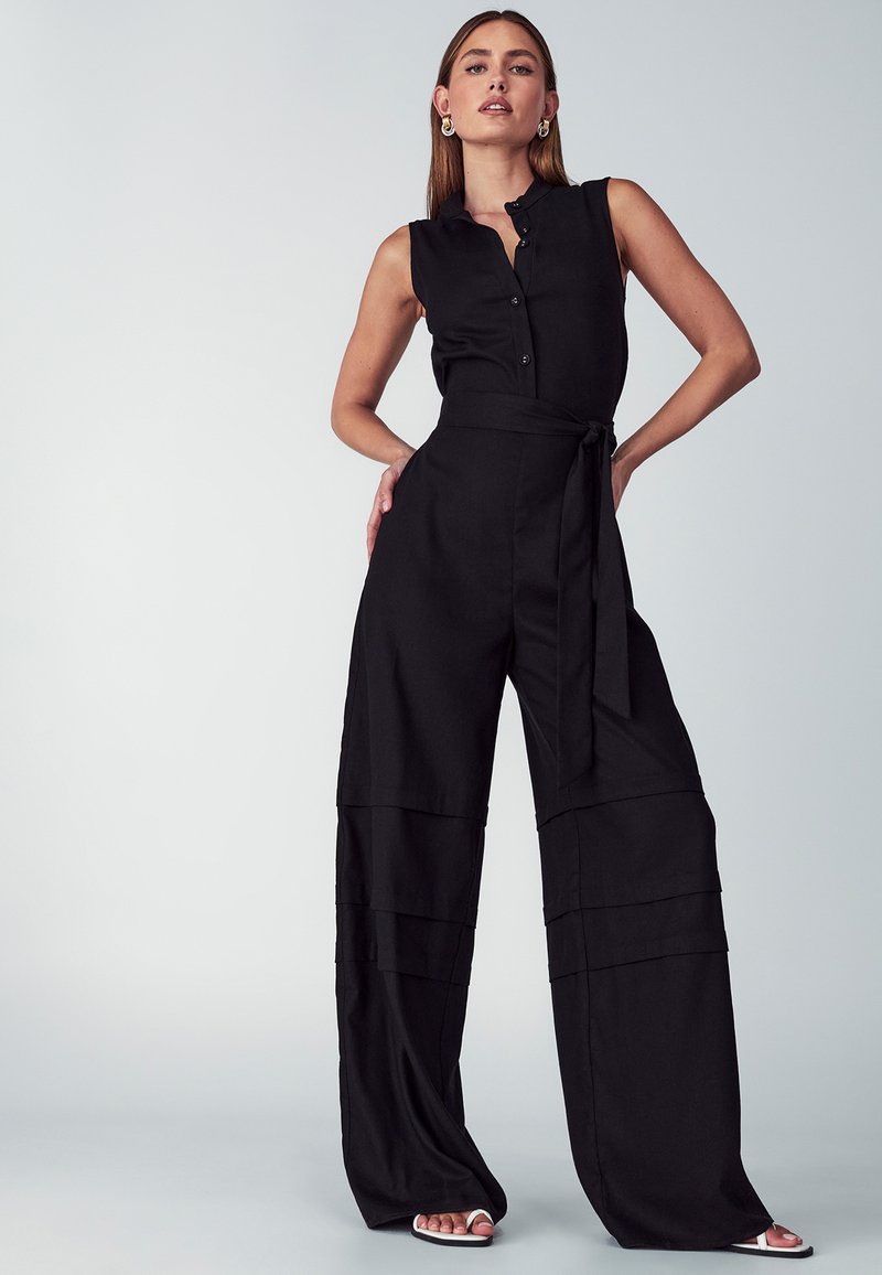 Črni kombinezon brez rokavov z gumbi spredaj in zavezanim pasom. Ima široke, ležerne hlačnice z večplastnimi odseki. Gladek tekstura.