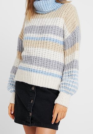 Femme portant un pull à col roulé en maille rayée beige, gris, blanc et bleu, associé à une mini-jupe noire avec des boutons sur le devant.