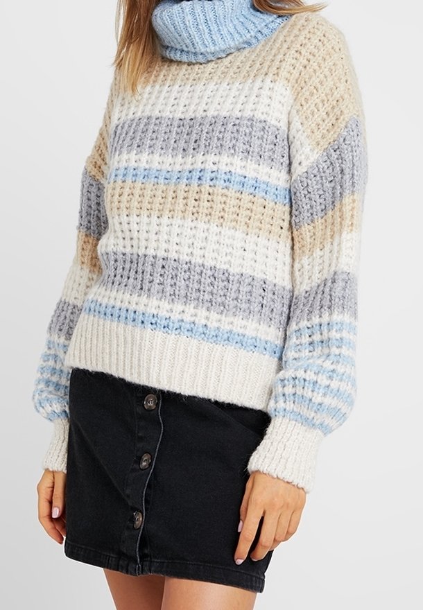 Femme portant un pull à col roulé en maille rayée beige, gris, blanc et bleu, associé à une mini-jupe noire avec des boutons sur le devant.