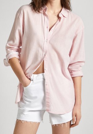 Kobieta ubrana w jasnoróżową koszulę na guziki z podwiniętymi rękawami i białe, postrzępione szorty jeansowe, z jedną ręką w kieszeni, stojąca na jednolitym tle.