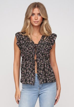 Frau mit langen blonden Haaren trägt schwarze ärmellose Bluse mit Blumenmuster und Rüschen sowie hellblaue Jeans vor einem schlichten hellen Hintergrund.