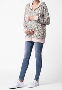 Felpa premaman con motivo floreale in verde e rosa, con zip frontale, cappuccio e polsini a costine. Indossata con jeans skinny e scarpe da ginnastica bianche.