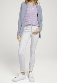 Ljusgrå cardigan över en lila randig skjorta med vit text, i kombination med vita slim-fit jeans och vita sneakers. Enkelt mönster och färger.