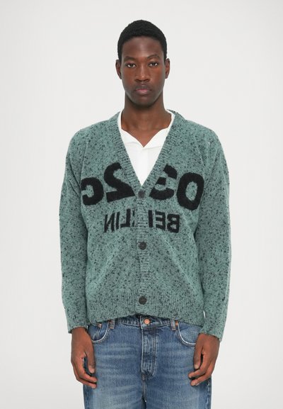 Grønt pletmønstret cardigan med V-hals, store sorte bogstaver, knaplukning og ribbede ærmer, parret med blå jeans.