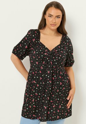 Vrouw in zwarte bloemenjurk met pofmouwen en geplooide taille, poseert met hand op heup tegen een effen achtergrond.