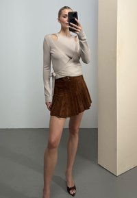 Γυναίκα βγάζει selfie στον καθρέφτη φορώντας μπεζ πουλόβερ τύπου wrap με άνοιγμα στους ώμους, καφέ πλισέ μίνι φούστα και σκούρα καφέ ανοιχτά παπούτσια.