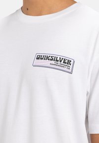 Camiseta blanca de algodón con un parche de logo negro y morado que dice "Quiksilver THE ORIGINAL BOARDSHORT CO." encima del lado izquierdo del pecho.