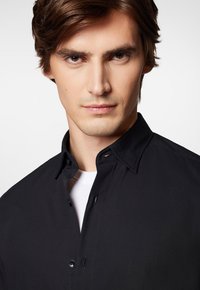 Czarna koszula zapinana na guziki z kołnierzykiem rozłożonym, z subtelnymi detalami w postaci przeszyć i gładką fakturą. Noszona na białej podkoszulkę.