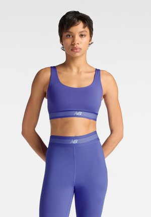 Person steht mit dem Gesicht nach vorne und trägt einen passenden lila Sport-BH und Leggings mit weißen gestreiften Taillenbändern und Markenlogo.