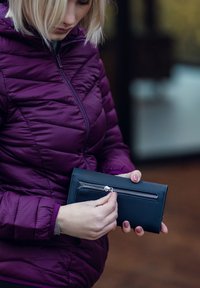 Black leather wallet with a zippered compartment, held by a person in a purple puffer jacket, featuring a smooth texture and metal hardware.