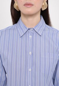 Femme portant des boucles d'oreilles abstraites dorées et une chemise à rayures verticales bleu et blanc avec une poche poitrine.