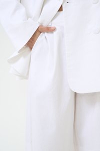 Blazer en lin blanc avec une coupe décontractée et des manches larges, associé à un pantalon en lin à jambes larges avec une poche latérale. Tissu doux et texturé.