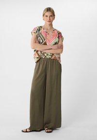 Bunt gemusterte Bluse mit kurzen Ärmeln, kombiniert mit leichten olivgrünen, weit geschnittenen Hosen und schlichten schwarzen Sandalen. Glatte Materialien sind durchgehend vorhanden.