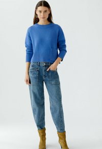 Blauer Strickpullover mit ribbeligem Saum und Tasche, kombiniert mit lockeren hellblauen Jeans mit mehreren Taschen und hellbraunen Ankle Boots.