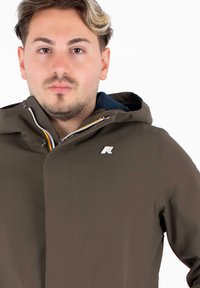Giacca impermeabile marrone con colletto alto, chiusura frontale zip e logo sul petto. Il materiale ha una texture liscia e raffinata.