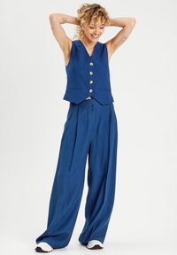 Gilet bleu avec cinq boutons et deux poches, associé à un pantalon bleu taille haute et ample. Tissu léger, texture lisse.