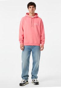 Felpa rosa con tasca frontale e cappuccio regolabile, abbinata a jeans azzurri chiari, leggermente strappati e sneakers bianche e nere.