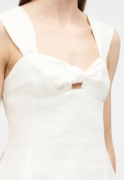 Λευκό αμάνικο φόρεμα με φαρδιές τιράντες και λεπτομέρεια κόμπου μπροστά που δημιουργεί μια μικρή κεντρική ανοίγμα, φορεμένο από άτομο.