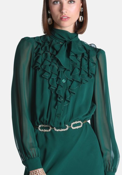 Femme portant un chemisier vert foncé à volants avec des manches transparentes, un nœud au cou, une ceinture chaîne scintillante et de grandes boucles d’oreilles en cristal.