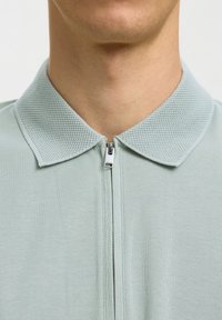 Gros plan sur le cou d'une personne portant un polo texturé bleu clair avec une fermeture éclair frontale partiellement fermée.