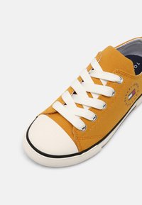Gul canvas sneakers med vita snören, gummitåhätta och svart sula. Har en logotypapplikation på sidan med cirkulära designaccenter.