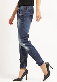 Jeans ajustados de mezclilla azul con acabado desgastado, detalles deteriorados y cinco bolsillos, combinados con zapatos negros de tacón alto.