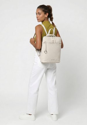 Nainen, jolla on punottu poninhäntä, valkoiset housut ja oliivinvihreä paita, kantaa beigen väristä suorakaiteen muotoista reppua, jossa on etutasku vetoketjulla.