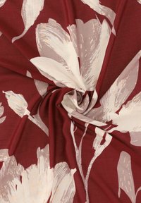 Czerwony materiał z dużymi, jasnobeżowymi wzorami kwiatowymi. Tkanina jest gładka i lekko układa się w fałdy, ukazując miękkie, teksturowane wykończenie.