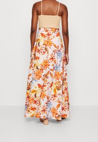 Žena v béžovém crop topu a dlouhé, splývavé sukni s květinovým vzorem v oranžové, žluté a modré barvě, stojící na jednobarevném pozadí.
