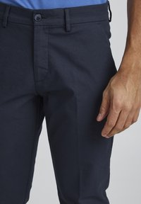 Pantaloni blu navy con texture liscia, frontale piatto, due tasche e chiusura con bottone; mano visibile appoggiata sul tessuto.
