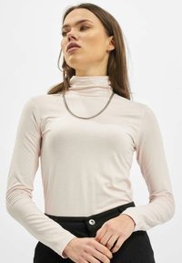 Gaišrozā garām piedurknēm turtlenecks blūze ar gludu auduma tekstūru, kurai pievienota sudraba ķēdes kaklarota un melnas augsto jostasvietu bikses.