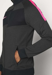 Person in einer dunkelgrauen Zip-Up-Jacke mit schwarzem Brustpanel und einem leuchtend pinken Streifen entlang der Schulter, mit der Hand in der Seitentasche.