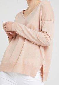 Jasnoróżowy sweter wykonany z miękkiej tkaniny, z dekoltem w serek, długimi rękawami i rozcięciami po bokach. Wykończony ściągaczami na mankietach i u dołu.