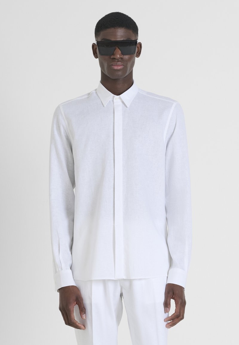 Camicia bianca a maniche lunghe realizzata in tessuto leggero, con chiusura a bottoni, colletto puntato e polsini. Gli occhiali da sole neri completano il look.