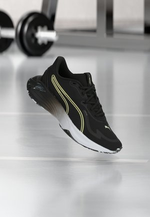 Juoda ir balta sportinė kedų pora su geltonais akcentais, sklendžianti virš atspindinčios sporto salės grindų, fone neryškios sporto salės svoriai.