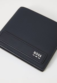 Jūras zila maku ar teksturētu virsmu, kas satur gluda ādas akcentu un metalizētu "BOSS" logotipu. Kompakta taisnstūra forma.