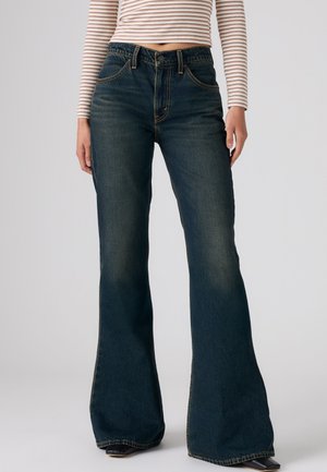 Personne portant un jean évasé taille haute bleu foncé et un top rayé court à manches longues, debout devant un fond uni.