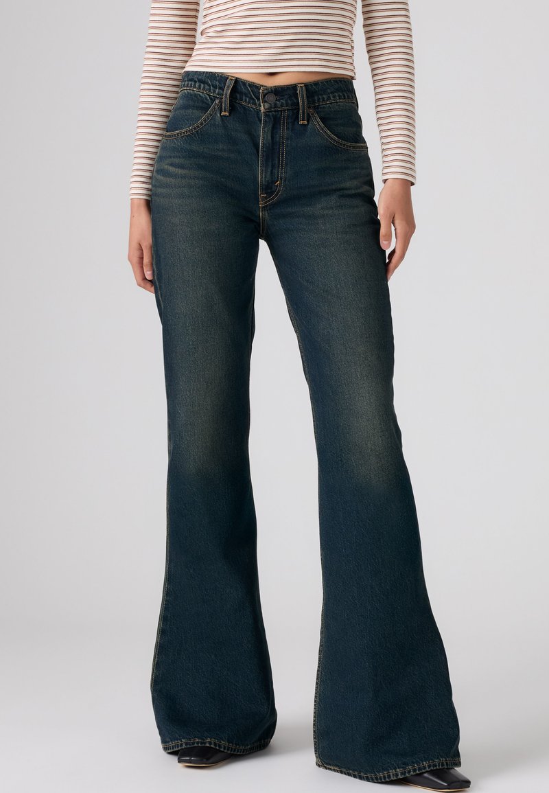 Personne portant un jean évasé taille haute bleu foncé et un top rayé court à manches longues, debout devant un fond uni.