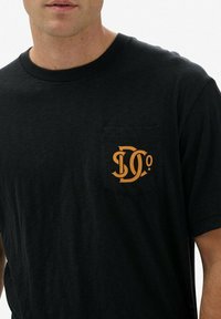 Camiseta de algodón negra con un bolsillo frontal y letras gráficas naranjas. Diseño clásico de cuello redondo con mangas cortas y un ajuste relajado.