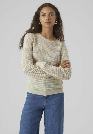 Pull léger beige avec un motif en tricot texturé, des manches longues et des poignets côtelés. Associé à un jean large bleu, sur un fond neutre.