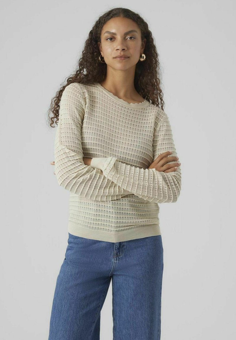 Maglione beige chiaro con pattern a maglia testurizzato, maniche lunghe e polsini a costine. Abbinato a pantaloni jeans a gamba larga blu, su uno sfondo neutro.