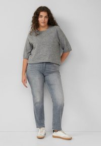 Kobieta z kręconymi włosami, ubrana w szary dzianinowy sweter, jasnoszare dżinsy i białe sneakersy, stoi przy gładkiej jasnoszarej ścianie.