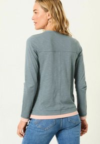 Langarmshirt aus hellgrauem Stoff, runder Ausschnitt, kontrastierende pinke Bündchen, strukturiertes Muster, taillierte Passform, getragen mit blauen Jeans.