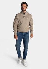 Beige gebreide trui met een hoge kraag en sluiting met twee knopen, gecombineerd met blauwe spijkerbroeken en witte sneakers. Eenvoudige, casual fit.